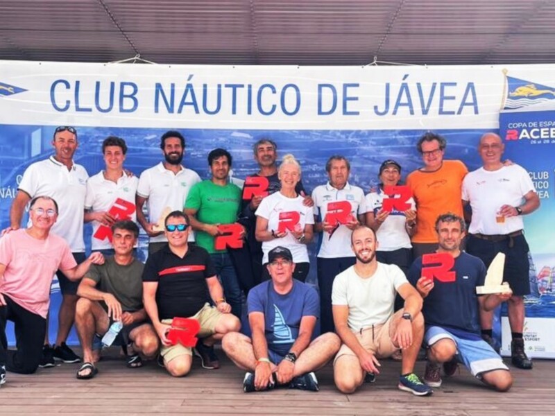 Equipo de personas posando para una foto frente a un cartel del Club Náutico de Jávea, con algunos portando insignias de la Copa de España de Racerboard.