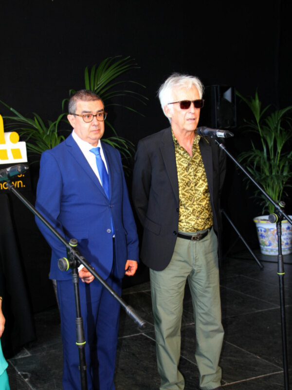 Dos hombres en una presentación formal, uno hablando en un micrófono y el otro escuchando.