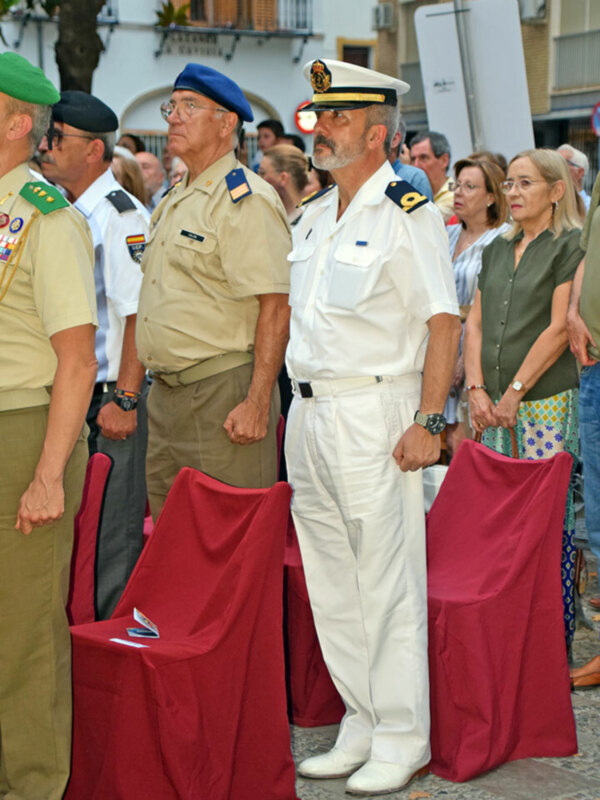 Un hombre en uniforme militar blanco y otro en uniforme beige, ambos con gorras, se encuentran en una ceremonia frente a una audiencia de personas vestidas formalmente. La escena ocurre en una calle con edificios y un árbol en el fondo.