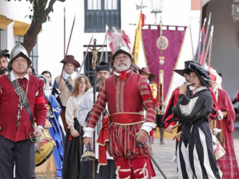 Reenactamiento histórico de una procesión medieval, con personajes vestidos con trajes rojos y negros, portando armas y banderas, en un entorno urbano con edificios blancos y azules.