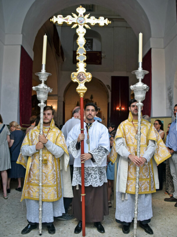 Oración religiosa en una iglesia con sacerdotes vestidos en vestimentas tradicionales, sosteniendo palos de procesión y velas. El ambiente es reverente, con arcos y velas encendidas en el fondo.