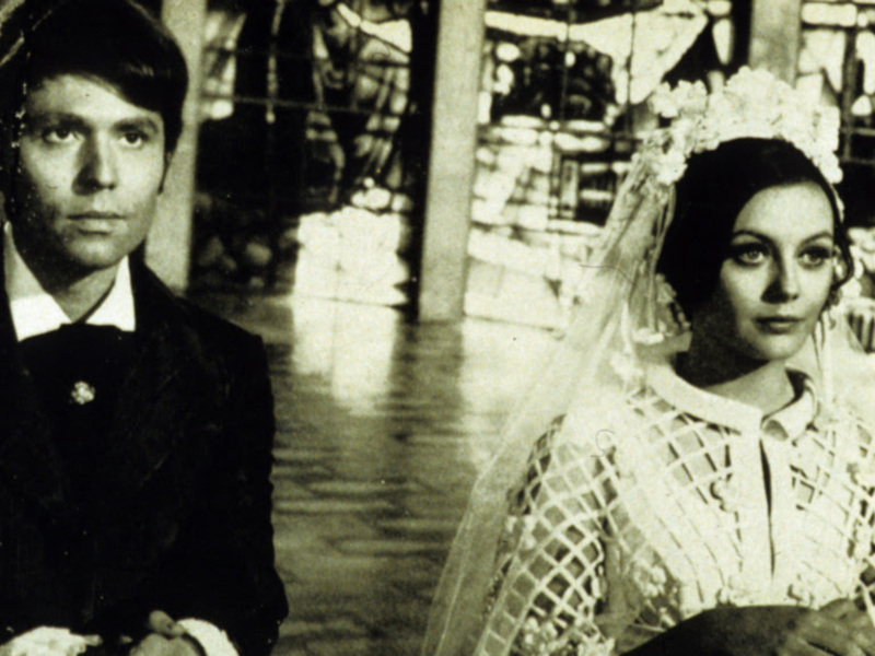 Una escena de una película en blanco y negro, con dos personajes en el centro de la imagen, uno de ellos con una vestimenta formal y el otro con un atuendo más informal y una corona en la cabeza.