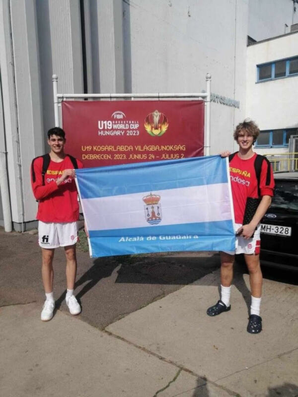 Dos jóvenes sostienen una bandera de Alcalá de Guadaira junto a una pancarta que anuncia el U19 FIBA World Cup en Hungría 2023. El fondo muestra un edificio blanco con una bandera roja y blanca.