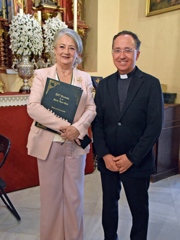 Una mujer en traje rosa sostiene un libro mientras posa junto a un hombre en traje negro. Ambos están en una sala con una mesa roja y flores blancas en el fondo.
