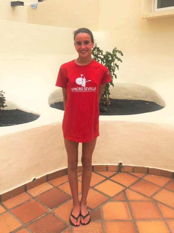 Una joven con una camiseta roja con el logo de Sincro Sevilla posa en un patio con baldosas de color naranja y un pequeño árbol en el fondo.