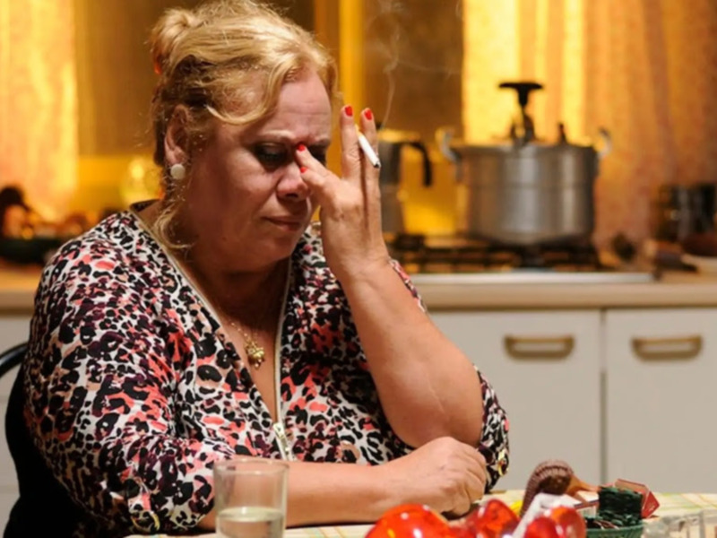 Una mujer en una cocina, fumando y tocándose el rostro, con un ambiente cálido y acogedor.