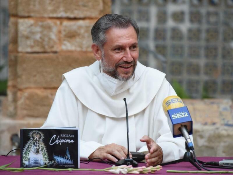 Monje hablando en rueda de prensa con un libro titulado 'Regla de Chipiona' en la mesa.