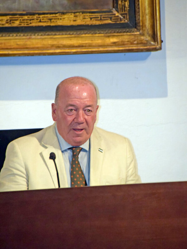 Hombre hablando en un evento formal en un podio con un marco dorado en el fondo.