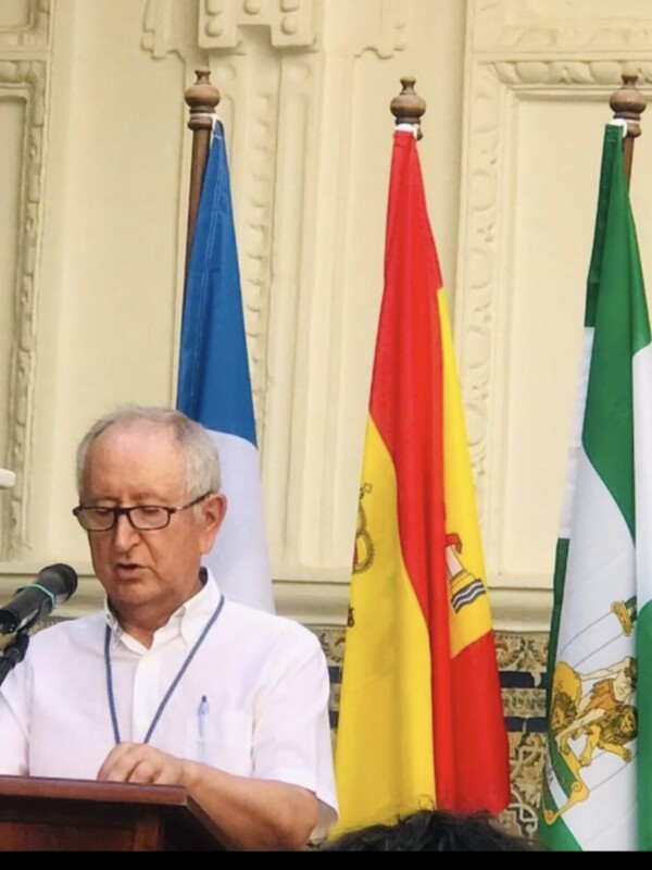 El fotógrafo capturó a un hombre en una ceremonia formal, posiblemente un evento político o diplomático. Se encuentra frente a un escenario con una silla y un microfono, rodeado por tres banderas: azul, roja con una estrella y verde. La decoración del lugar sugiere un ambiente oficial o institucional.