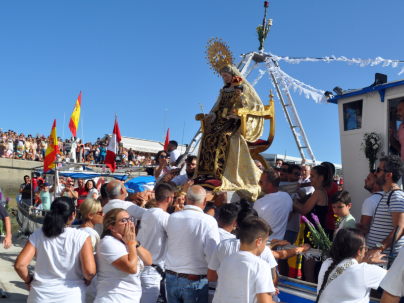Procesión marítima con la imagen de la Virgen del Carmen, rodeada por fieles en un puerto, bajo un cielo azul claro.