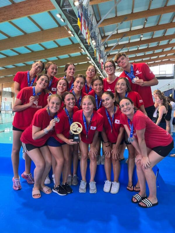 Equipo de waterpolo celebrando su victoria con medallas y trofeo en una piscina cubierta.