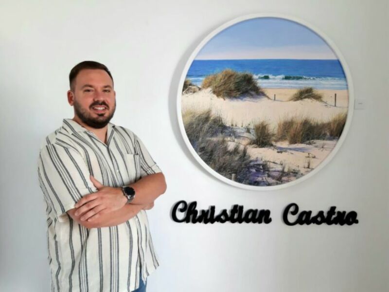 Christian Christian Castro posando junto a una imagen de una playa con dunas y un mar azul claro.