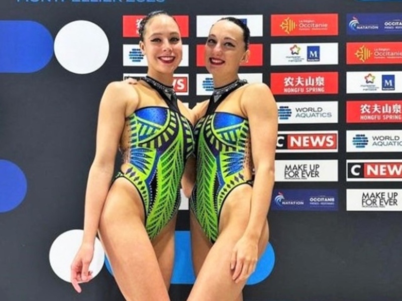 Duo de nadadoras sincronizadas posando en trajes de competencia con patrones vibrantes, frente a un fondo con logos de patrocinadores.