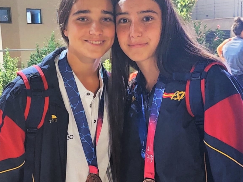 Dos personas sonrientes con medallas de oro en la boca, posando juntas en un entorno urbano con edificios y vegetación al fondo.
