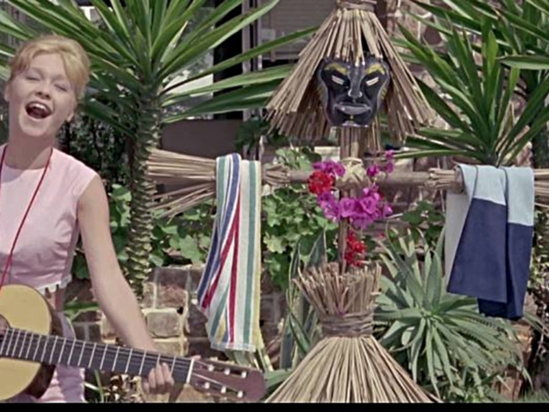 Una mujer toca la guitarra y canta en un entorno tropical con plantas y una máscara de madera en el fondo.