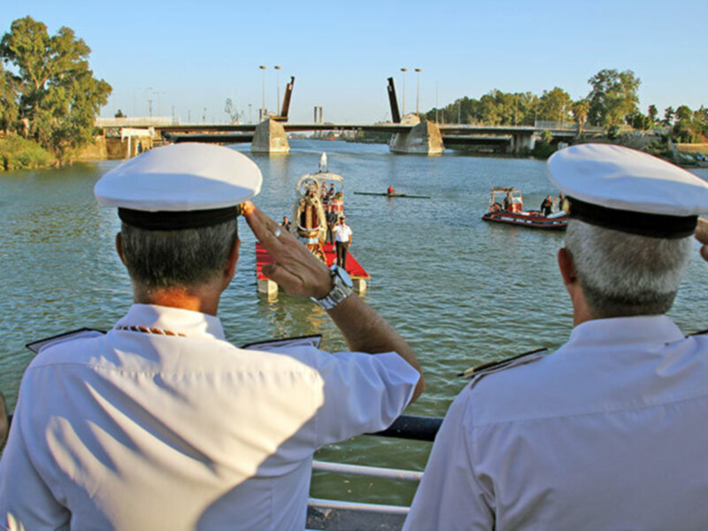 Oficiales de marina saludando en un evento en el agua, con barcos y puente al fondo.