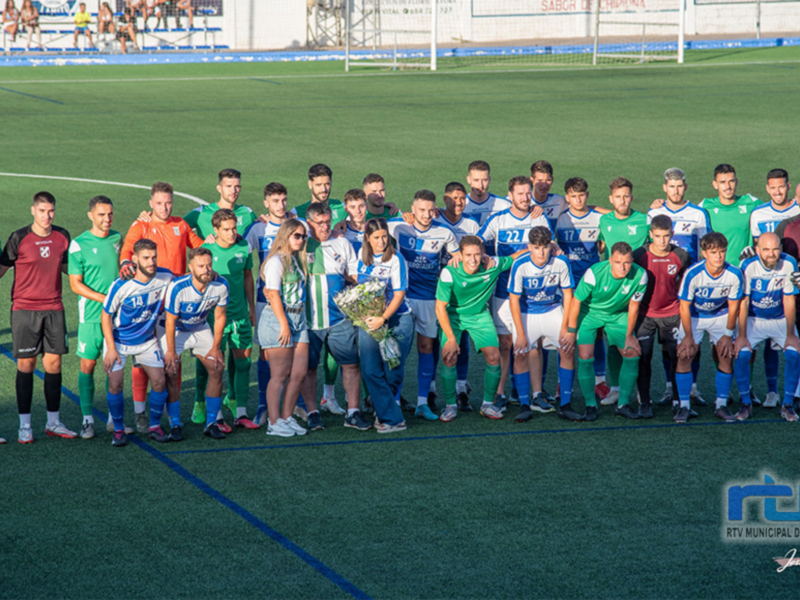 Equipo de fútbol posando en el campo de juego, con jugadores vestidos de verde y azul, y una mujer con un ramo de flores en el centro.