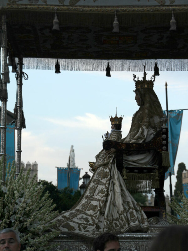 Estatua de una virgen con coronas y mantas en un evento ceremonial, rodeada por decoraciones y banderas azules.