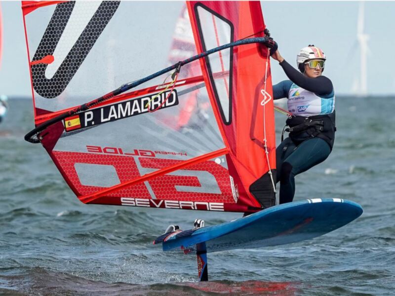 Competidor de windsurf en acción durante una competición, navegando con vela roja y nombre 'P. Lamadrid' visible.