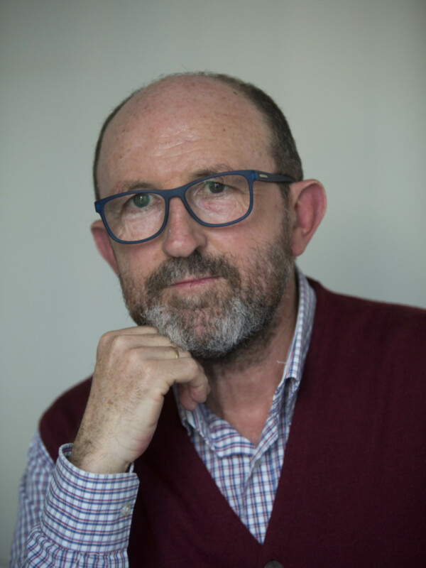 Retrato de hombre con gafas y barba, vestido con chaleco y camisa a cuadros, pensativo con la mano en la barbilla.