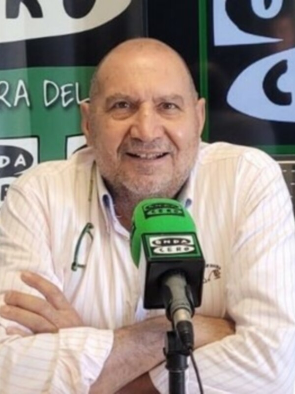Persona hablando en un estudio de radio con un micrófono de color verde y fondo con logos de emisoras.