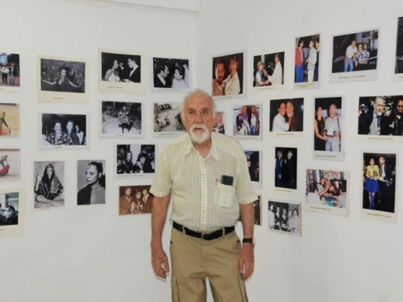 Un hombre mayor con barba blanca y camisa de rayas posa frente a una pared con varias fotos de personas en diferentes escenas.