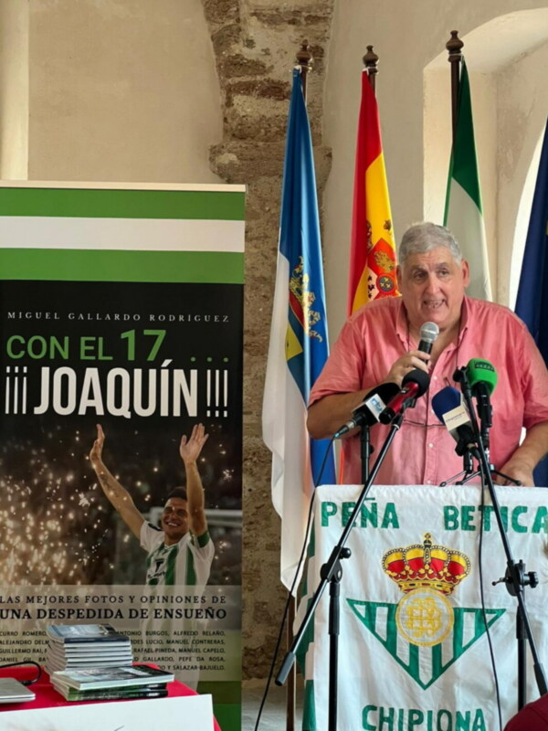 Entrevista con el presidente del Real Betis en un evento público, con banderas y un cartel promocional en el fondo.