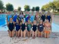 Una imagen de un grupo de 12 nadadoras en una piscina. Las nadadoras están vestidas con trajes de baño y posan juntas para la foto. La piscina está rodeada por una pista de atletismo con porterías y un césped. El cielo es claro y el ambiente es soleado.