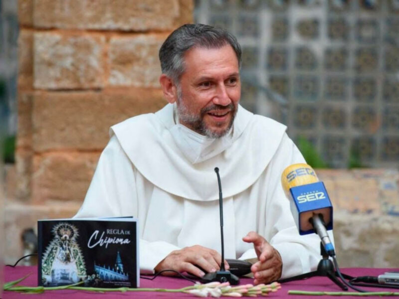 Monje hablando en una conferencia de prensa con un micrófono y un libro sobre la Virgen de Chipiona en la mesa.