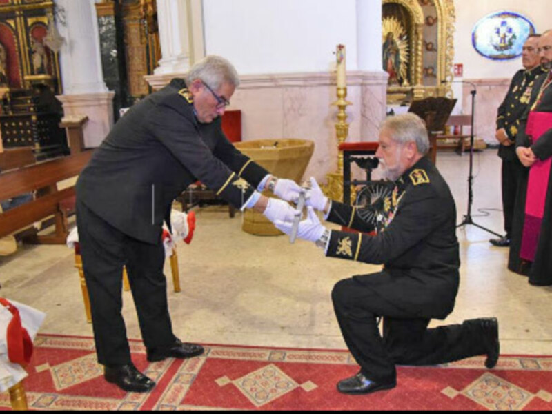 Un hombre en uniforme militar se inclina para recibir una insignia de un oficial de la policía, en un entorno formal con elementos decorativos y una persona en el fondo.
