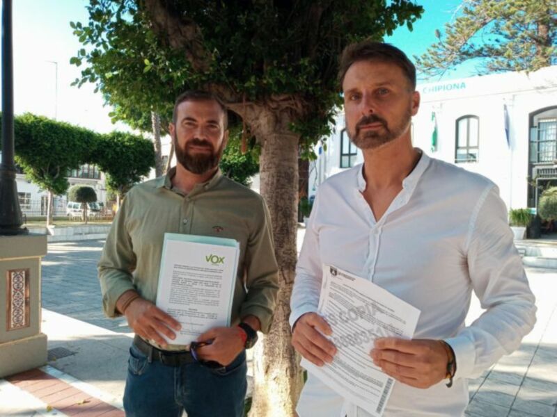 Dos hombres sostienen documentos en un entorno urbano con árboles y edificios en el fondo.