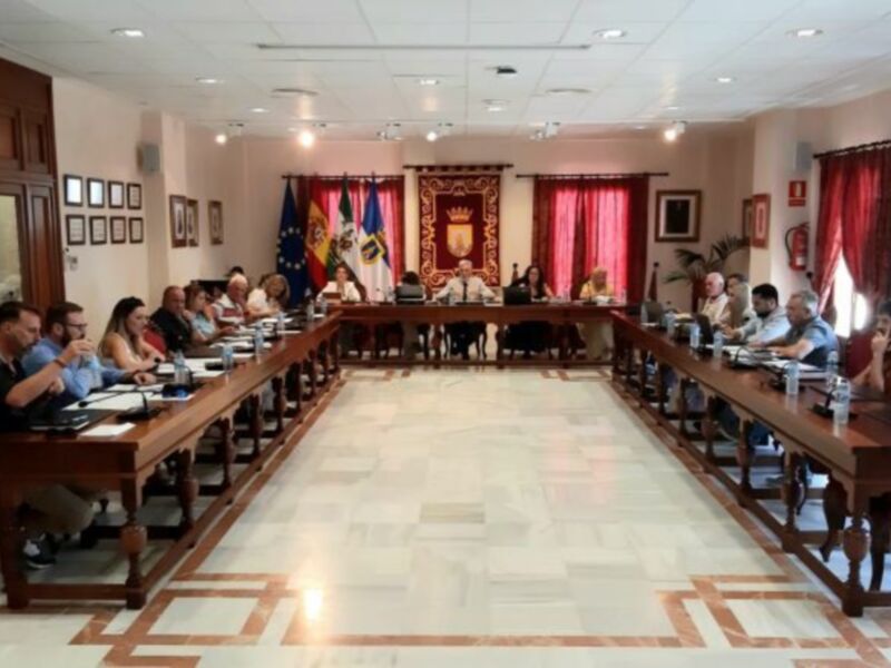 Sala de reuniones con una mesa larga y varias personas sentadas, decorada con banderas y un escudo heráldico en el fondo.