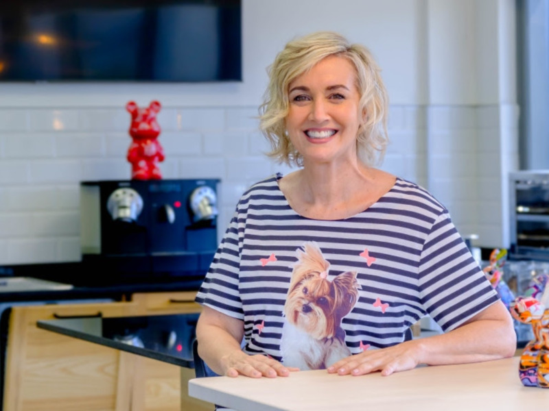 Una mujer sonríe en una cocina moderna, vistiendo una camiseta con una imagen de un perro. En el fondo, hay una estatua roja y un espacio bien iluminado.