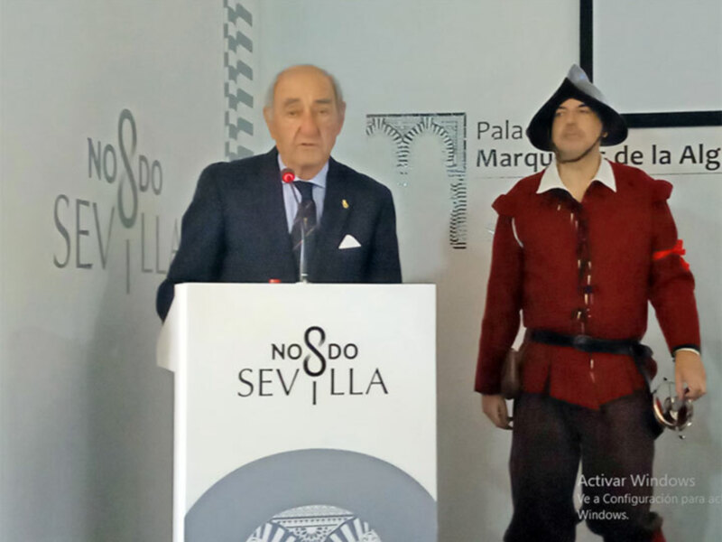 Un hombre en traje negro y corbata se dirige a un público, mientras otro individuo en traje rojo y sombrero se encuentra a su lado.
