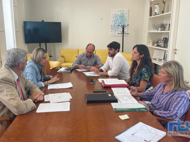 Un grupo de personas reunidas en una sala de conferencias, discutiendo y trabajando en documentos. La sala está equipada con una televisión, una mesa con documentos y teléfonos, y una silla amarilla en el fondo. La atmósfera es formal y productiva.