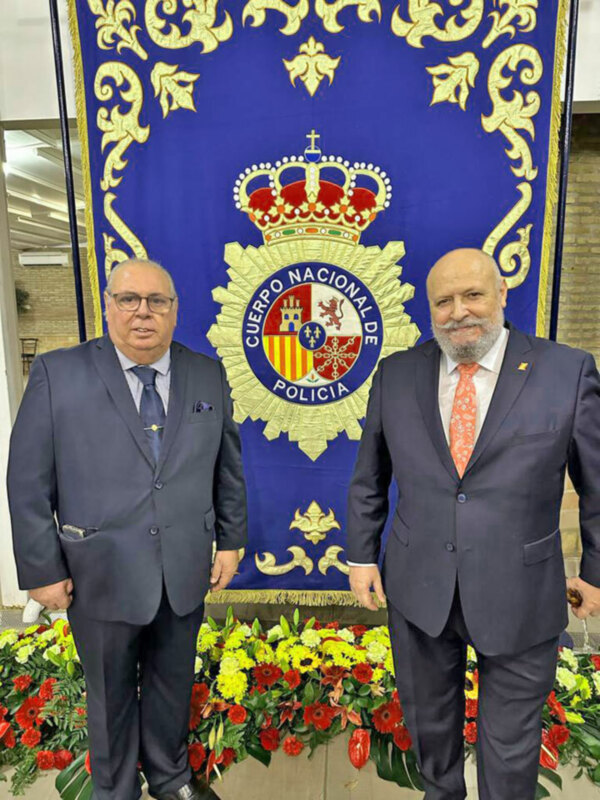 Dos hombres en trajes posan frente a un escudo de la Policía Nacional de España, con flores en el suelo.