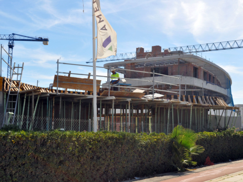 Estadio en construcción con una bandera AME en el fondo y trabajadores en la obra.