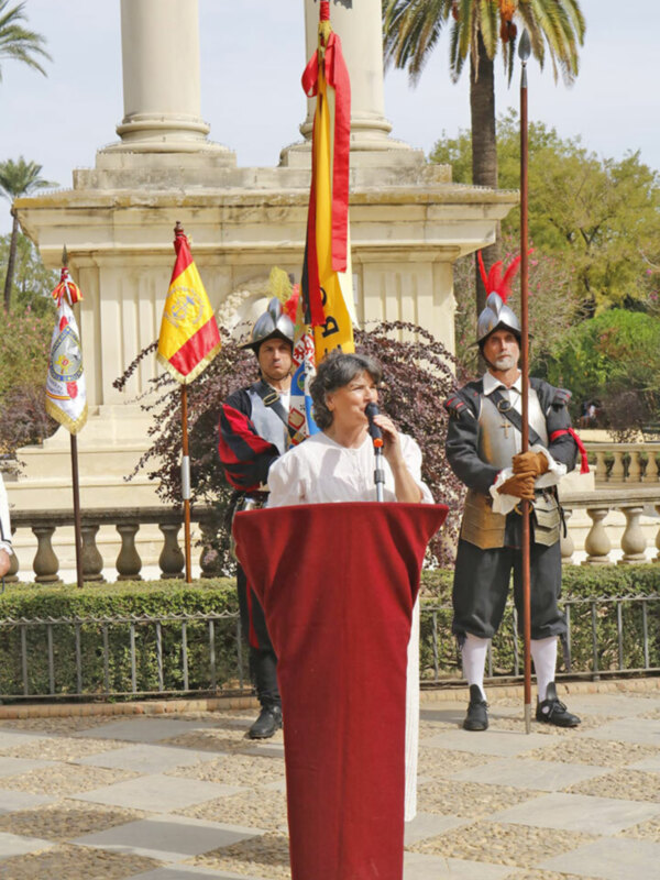 Una mujer en un podio habla mientras dos soldados históricos con cascos y banderas españolas observan en un entorno ceremonial.