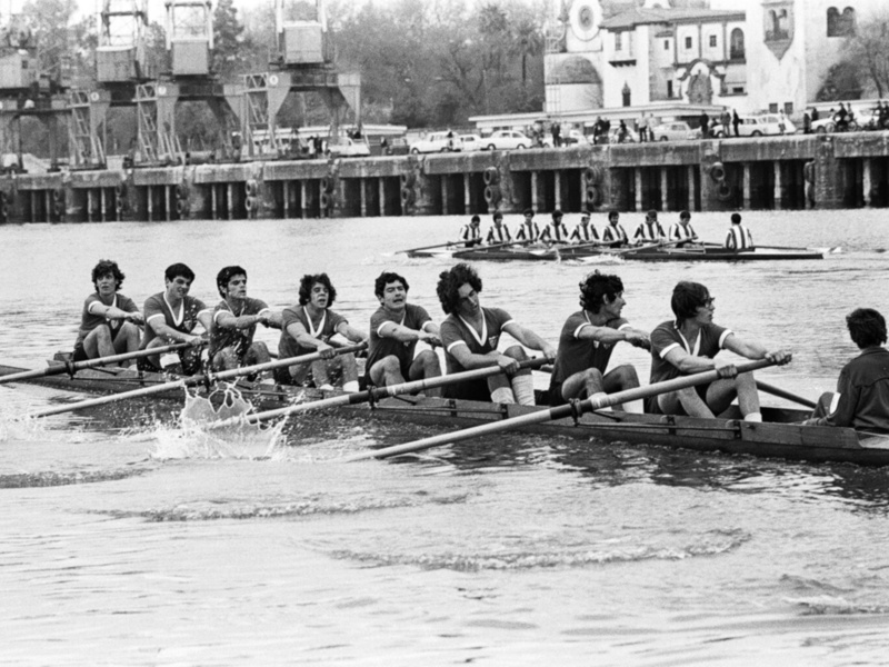 Equipo de remo en acción durante una competición en el río, con espectadores y estructuras portuarias al fondo.