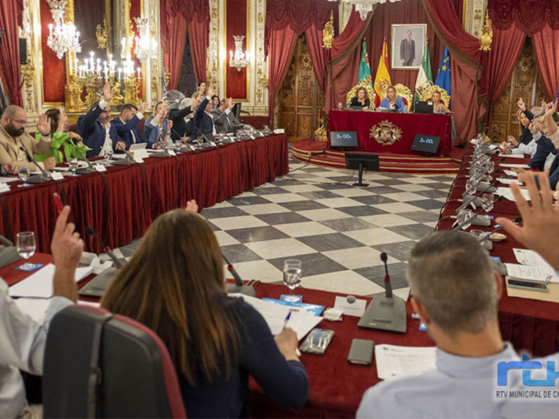 Reunión de autoridades en un salón decorado con cortinas rojas y lámparas de cristal, con participantes levantando las manos para votar.