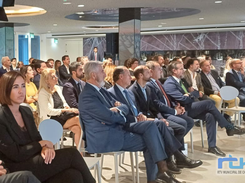 Asistentes de una conferencia en una sala moderna, con personas sentadas en sillas blancas y vestidos formales. El ambiente es formal y profesional, con un fondo que parece ser una pantalla de televisión.