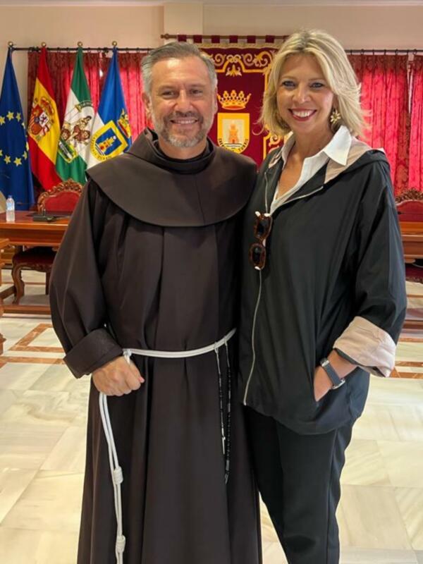 Un monje en hábito y una mujer en traje, posando frente a banderas europeas y nacionales, posiblemente en un evento oficial.
