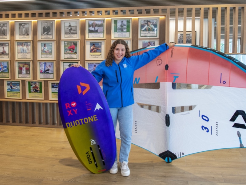 Una persona sostiene un kite surf y una tabla de surf en un entorno moderno con fotos de deportistas enmarcadas en la pared.