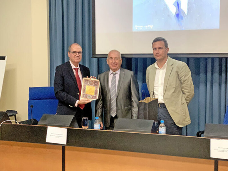 Tres hombres en trajes posando con un libro en una sala de conferencias.