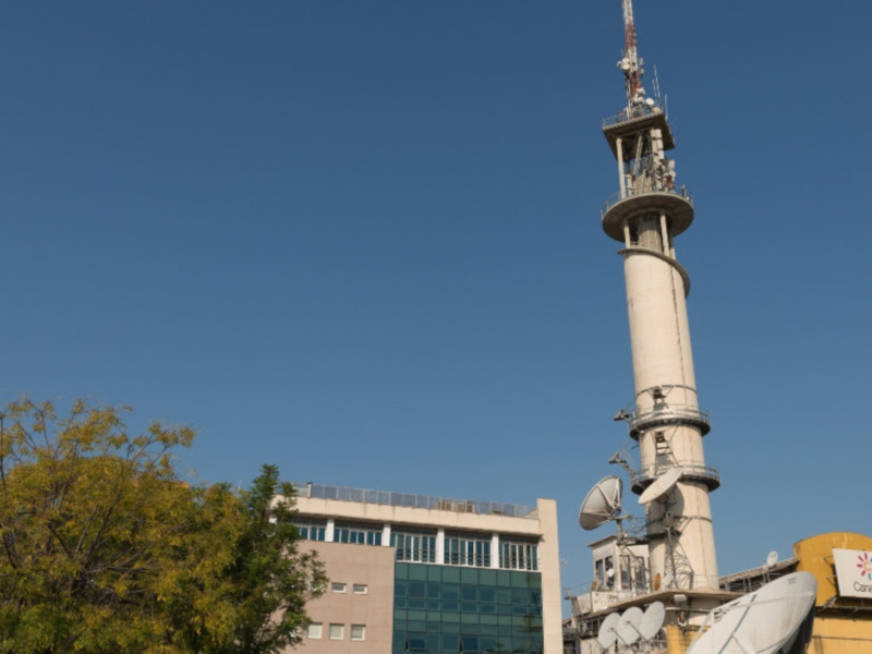 Torre de telecomunicaciones con antenas y satélites, frente a un edificio moderno bajo un cielo azul claro.