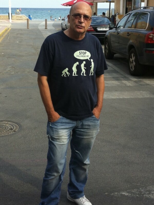 Man standing on a street with palm trees and a cafe in the background.