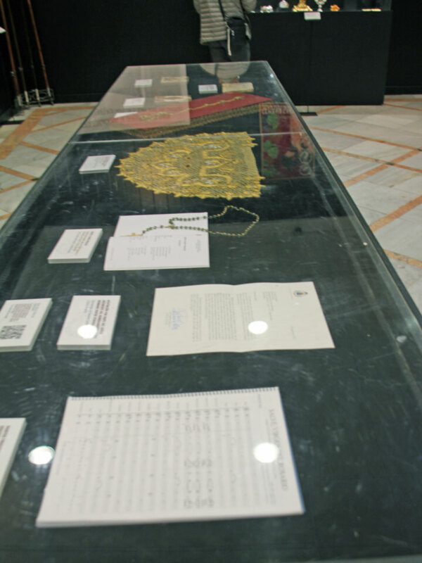 Exposición de objetos antiguos en una vitrina de cristal, con un fondo de madera y un suelo con patrones geométricos. Al fondo, se puede ver a una persona caminando. En la vitrina, hay documentos y una pieza de arte con un corazón dorado en el centro.