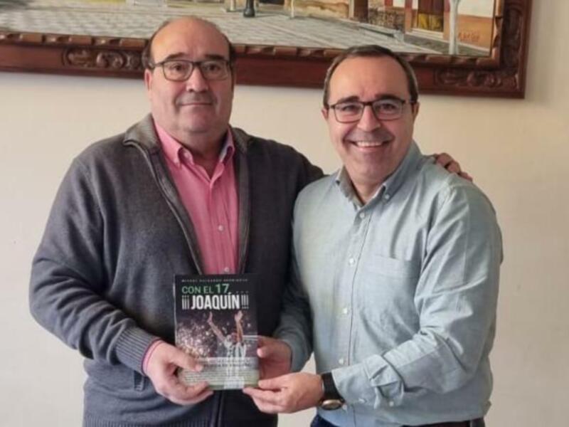 Dos hombres posan con un libro titulado 'Con el 17 Joaquín' en un entorno acogedor.