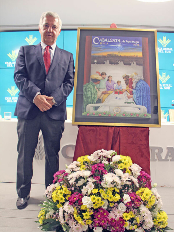 Un hombre en traje y corbata posa junto a un póster de la Cabalgata de Reyes Magos en Sevilla 2024, con un gran ramo de flores en el frente.