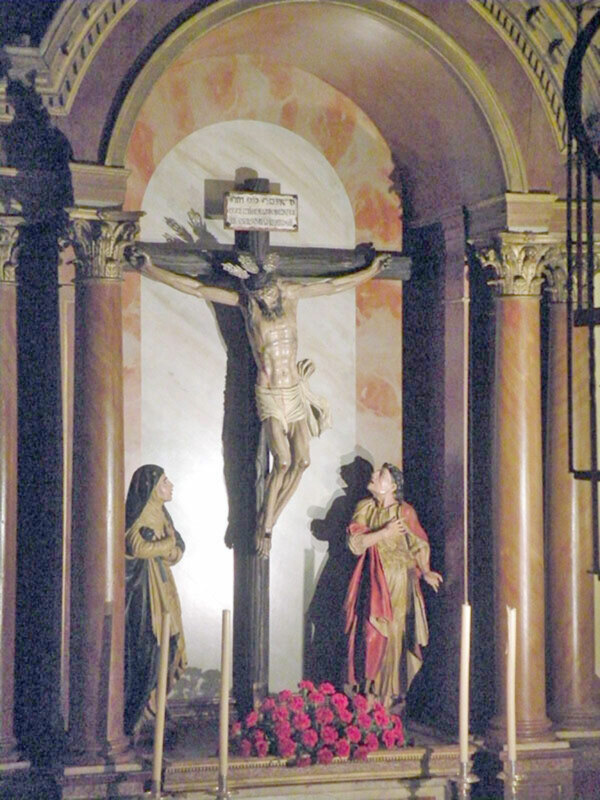 Escena religiosa con la figura de Cristo en la cruz, rodeado por dos mujeres en actitud de oración, en un interior de iglesia con arcos y columnas.